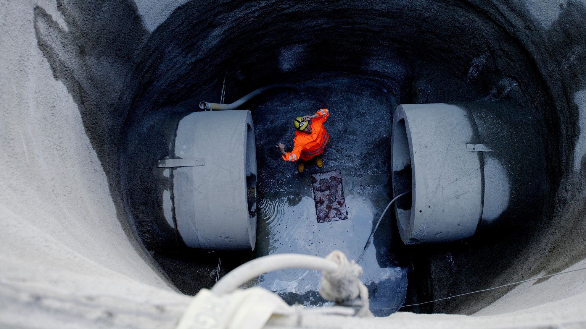 Blick von der Straße runter in eine tiefe Baugrube auf einen Kanalbaustelle, in deren Mitte zwischen zwei großen Abwasserkanalrohren ein Arbeiter in orangener Sicherheitsbekleidung steht.
