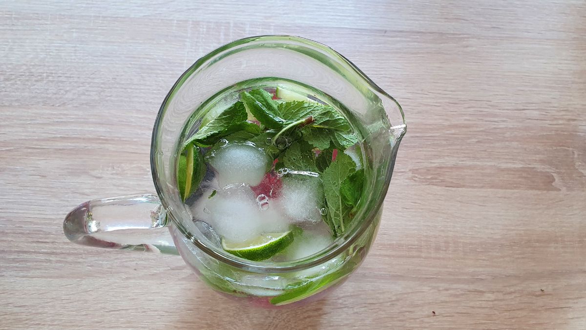 Blick in eine Glaskaraffe von oben, die Trinkwasser aus der Leitung, Eiswürfeln, Minze und Himbeeren gefüllt ist