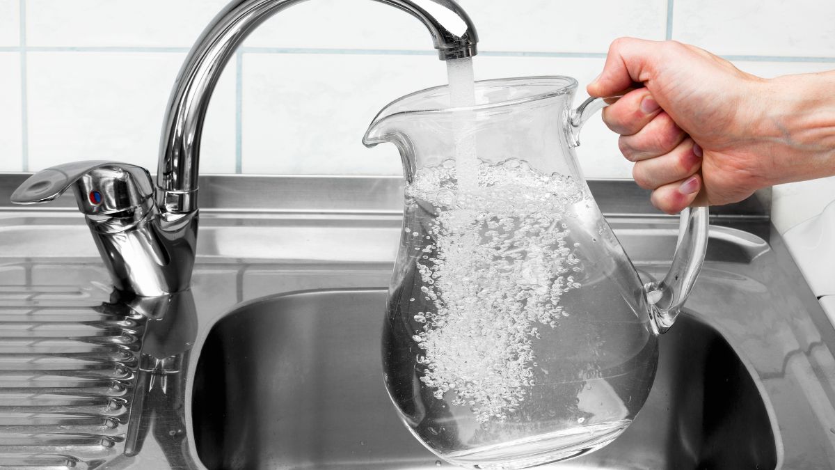 Eine Hand hält eine Glaskaraffe unter den Wasserhan in der Küche und lässt frisches Trinkwasser in die Karaffe laufen