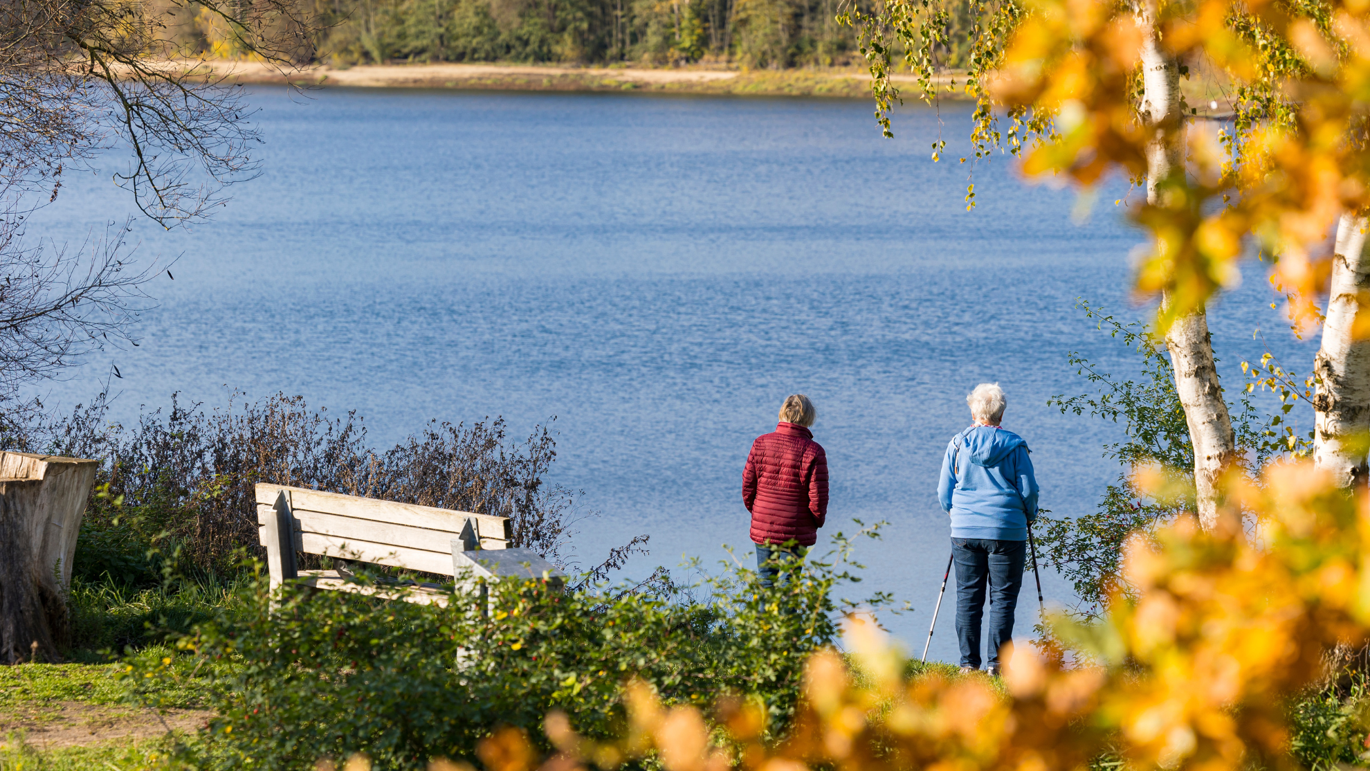 Blick auf die Talsperre Hullern im Herbst, im Vordergrund eine Bank und zwei Personen, die am Seeufer stehen und aufs Wasser schauen. © GELSENWASSER AG