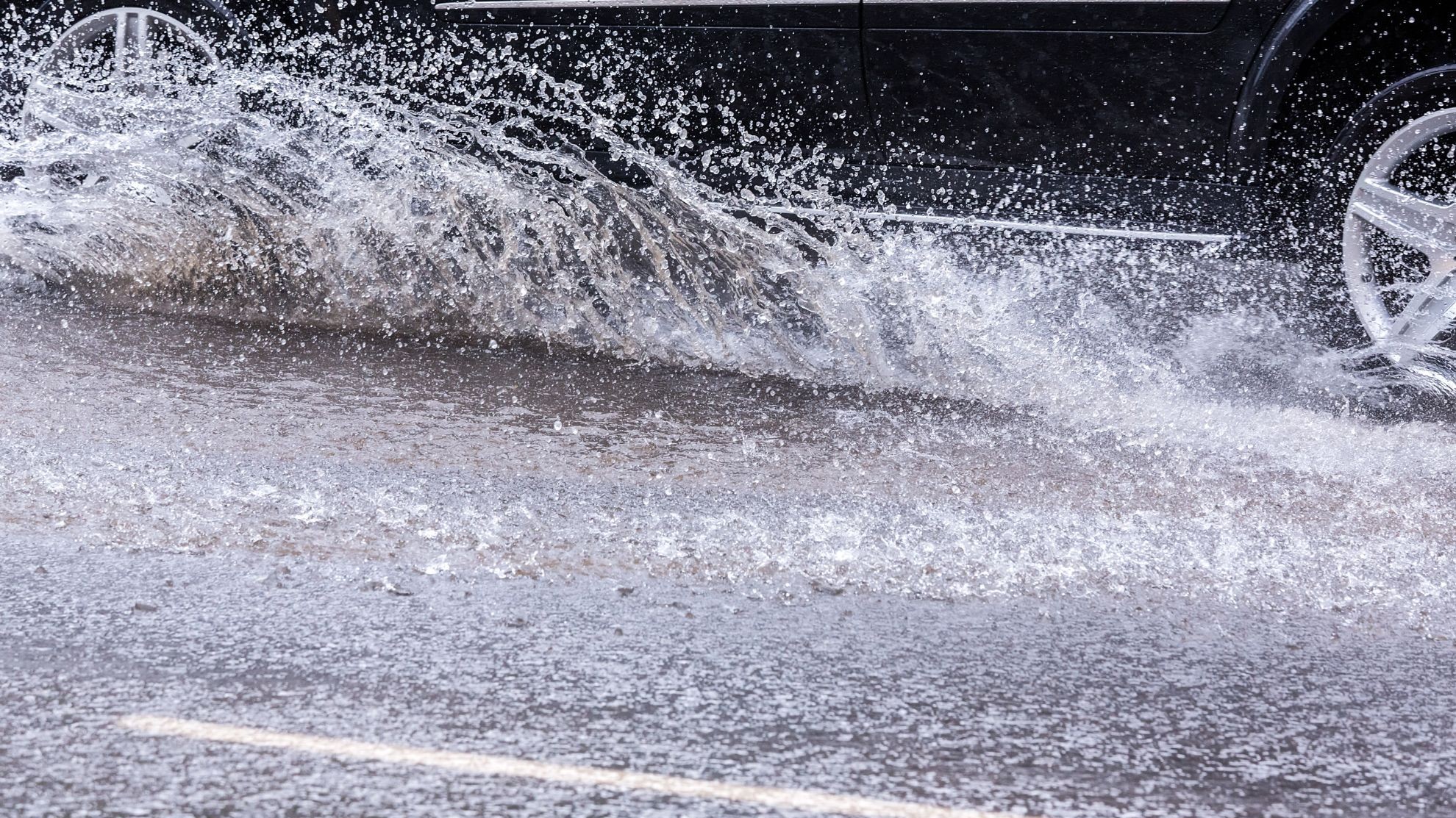 Ein Auto fährt im Starkregen über eine Straße und das Wasser spritzt hoch.