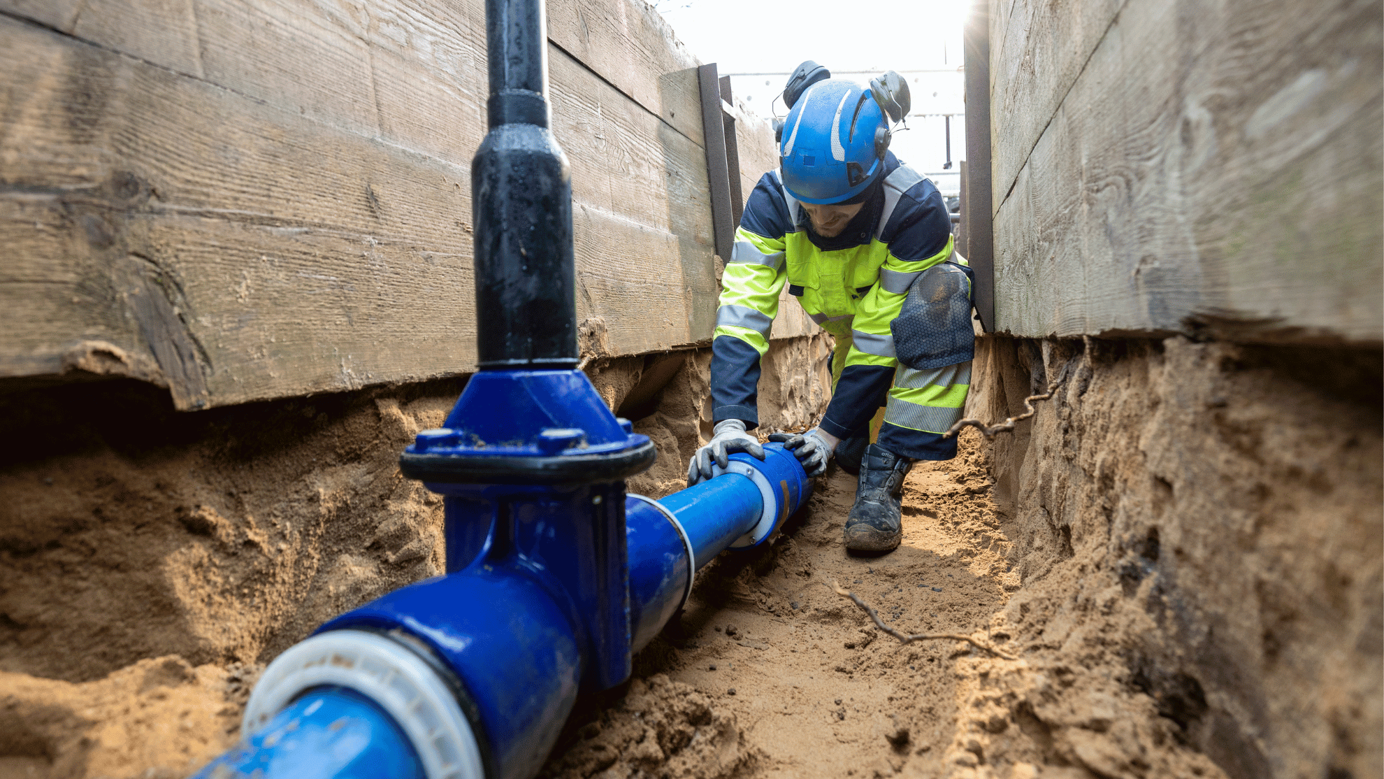 Ein Gelsenwasser-Mitarbeiter kniet in blau-gelber Schutzkleidung in einem Baugraben, vor sich eine größere Wasserleitung, die neu verlegt wurde mit einem Hydranten.